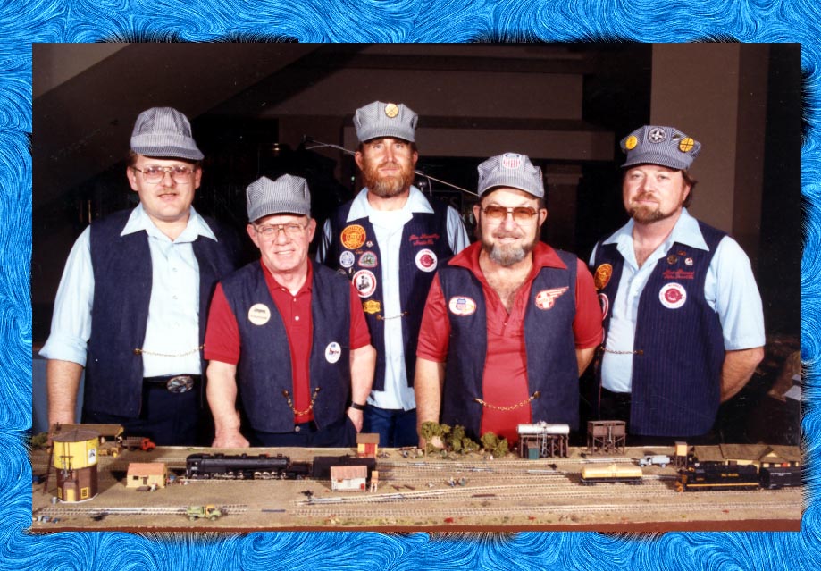 Five men in matching vests and conductor caps pose proudly behind a model train display, showcasing their passion for model railroading.