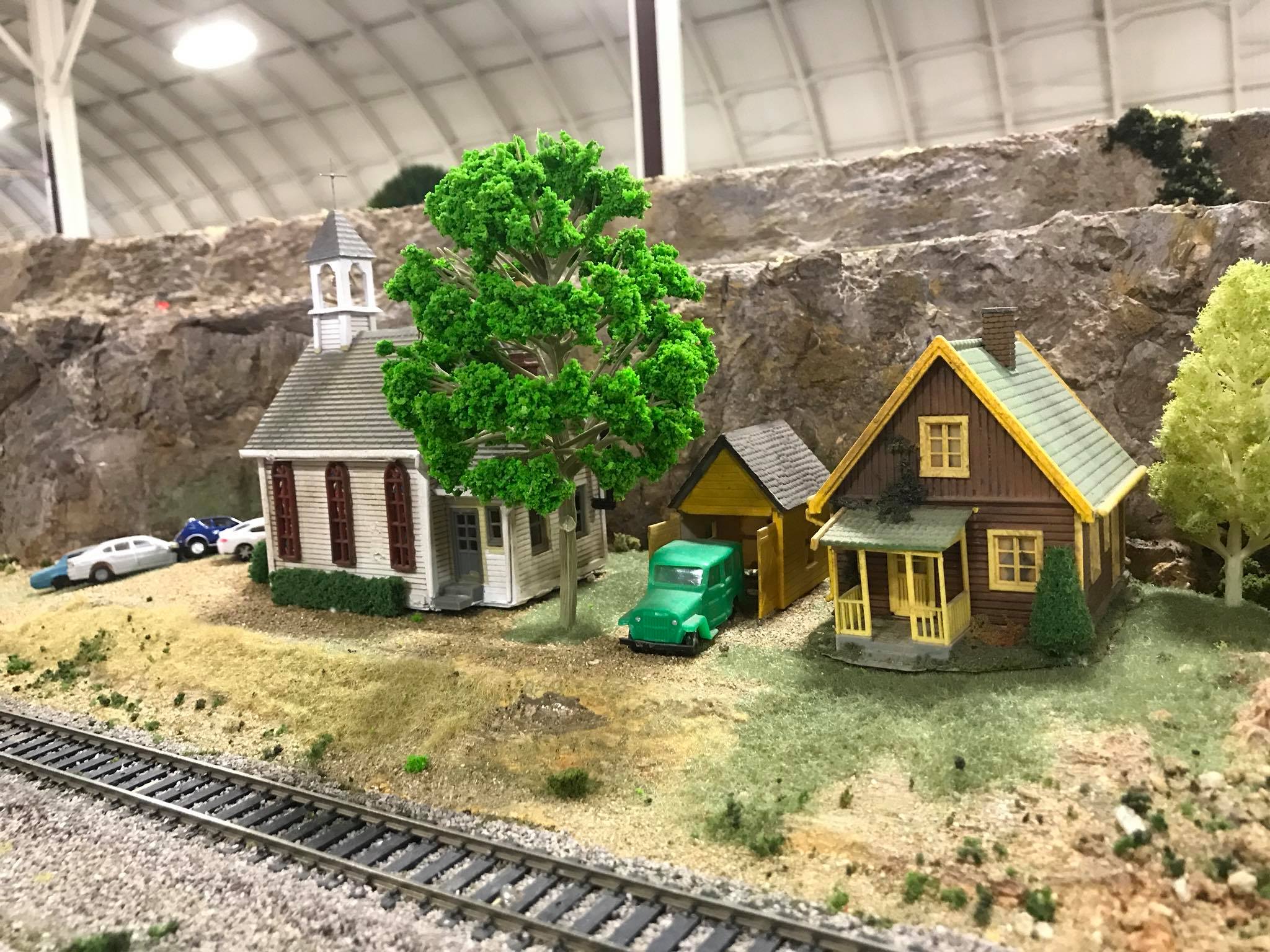 Miniature scene featuring a church and two houses near a railway track, showcasing detailed model landscaping and vehicles. Ideal for train enthusiasts.