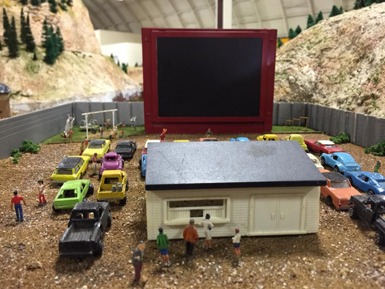 Miniature diorama featuring a drive-in movie setup with diverse toy cars, a small concession building, and tiny figures enjoying the scene.
