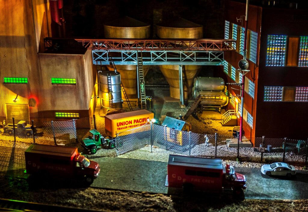 A detailed model scene depicting a Union Pacific freight yard, featuring trucks, silos, and a fire crew vehicle, illustrating industrial operations.