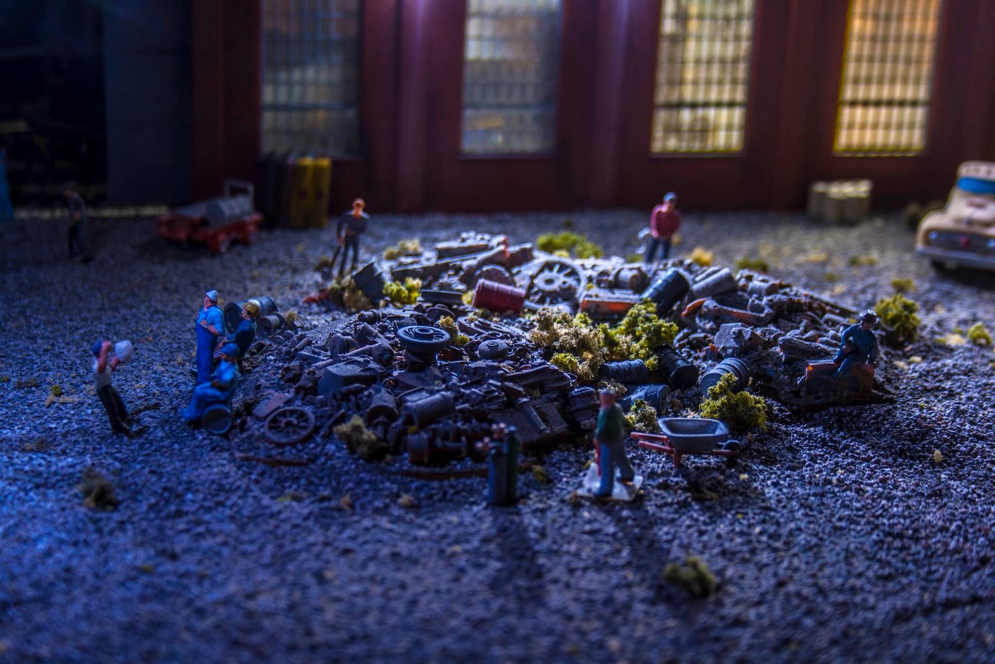 Miniature figures gather around a heap of junked machinery and debris, depicting a scene of salvage work in a cluttered workshop.