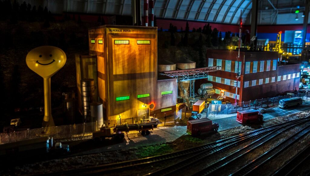 A detailed model of a paper factory featuring illuminated windows, a large smiling water tower, and various trucks, set on a miniature landscape.