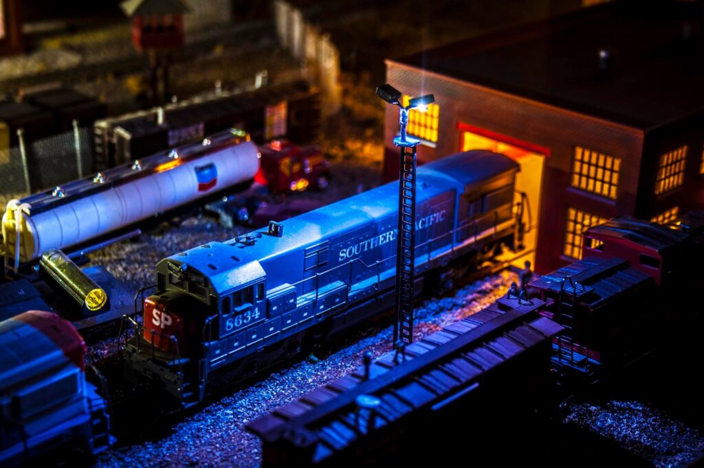 Blue model train labeled "Southern Pacific" is parked near a brightly lit building, surrounded by freight cars and a fuel tank, showcasing a miniature railway scene.