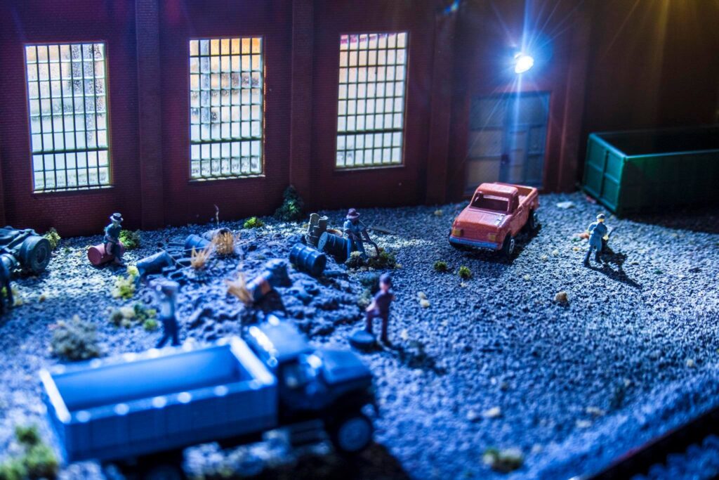Miniature scene depicting a realistic industrial yard with figures interacting among vehicles and debris, illuminated by overhead lights.
