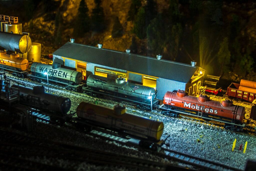 A model train yard at night featuring various tank cars, including "Warren" and "Mobilgas," alongside the "Grasshopper Oil Supply" building.