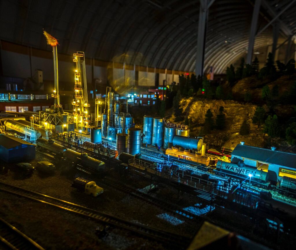 Illuminated model oil refinery scene with detailed tanks, rail cars, and equipment under a dramatic indoor setting, showcasing industrial scale.