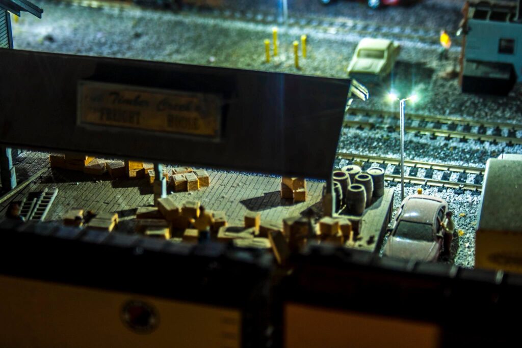 Detailed model train station scene featuring scattered crates, a parked car, and a figure interacting near a light post, showcasing railway logistics.
