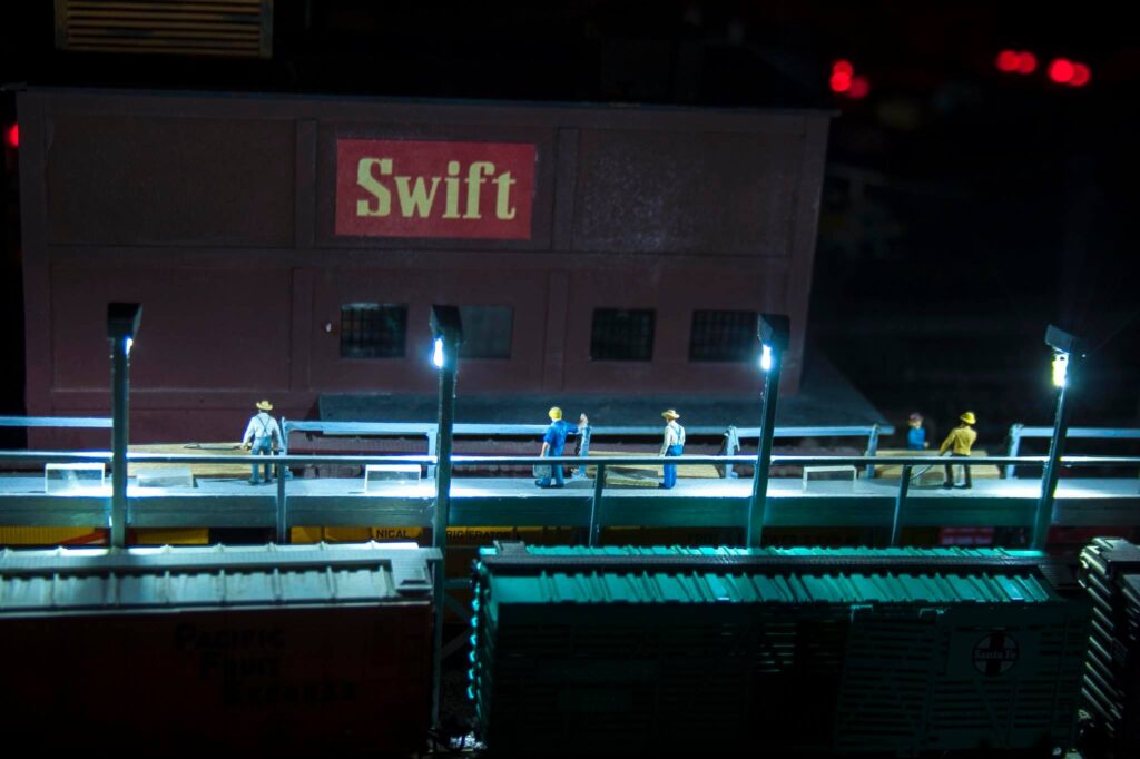 Miniature scene depicting figures on a platform beside freight trains, with a building labeled "Swift" illuminated at night.