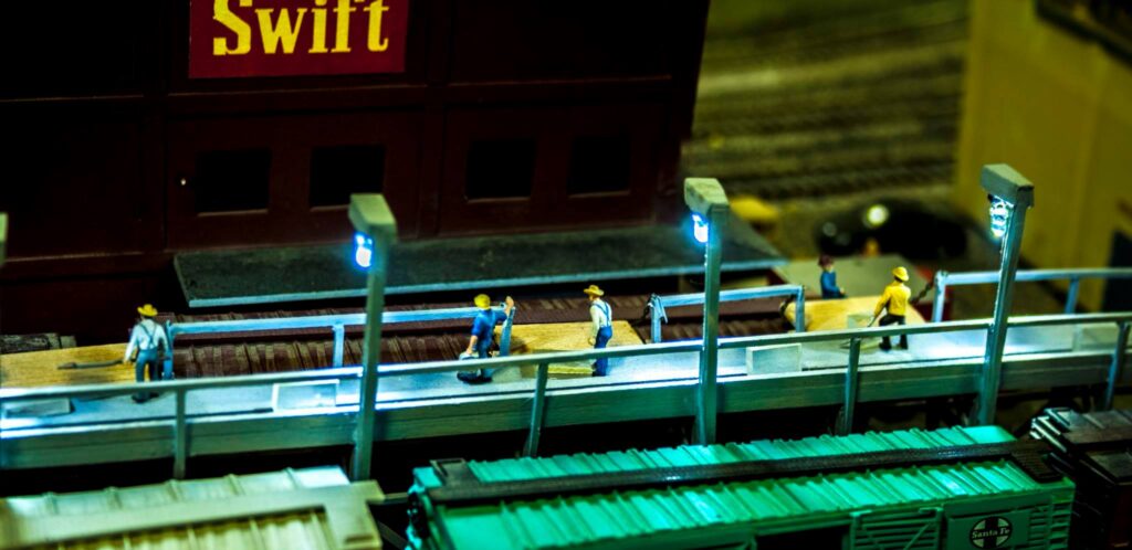 Six miniature figures working on a wooden platform in a model train setting, highlighting a freight loading scene at a Swift building.