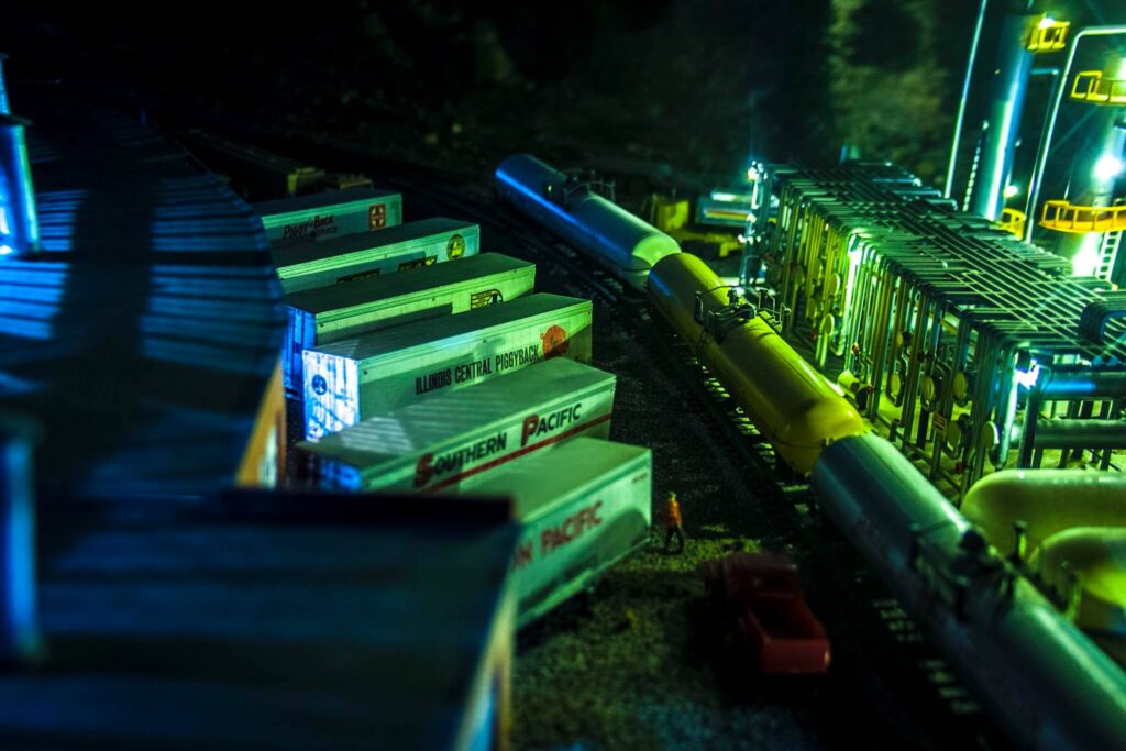 Model train scene featuring multiple freight cars and a yellow tank car at a miniature industrial yard, illuminated by green lights.