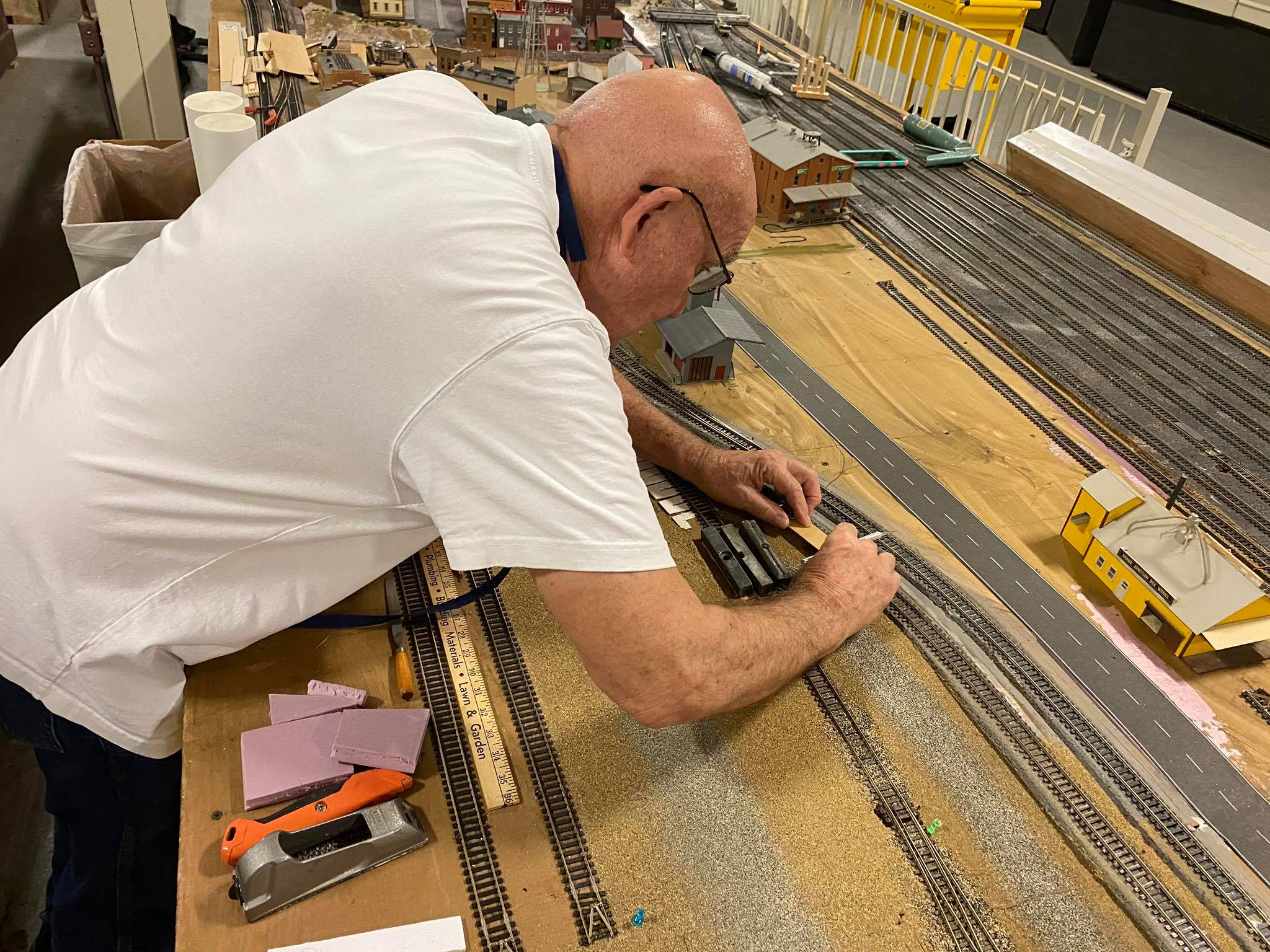 A model train enthusiast adjusts tracks on a detailed layout, surrounded by miniature buildings and scenery. The image captures hobby craftsmanship.