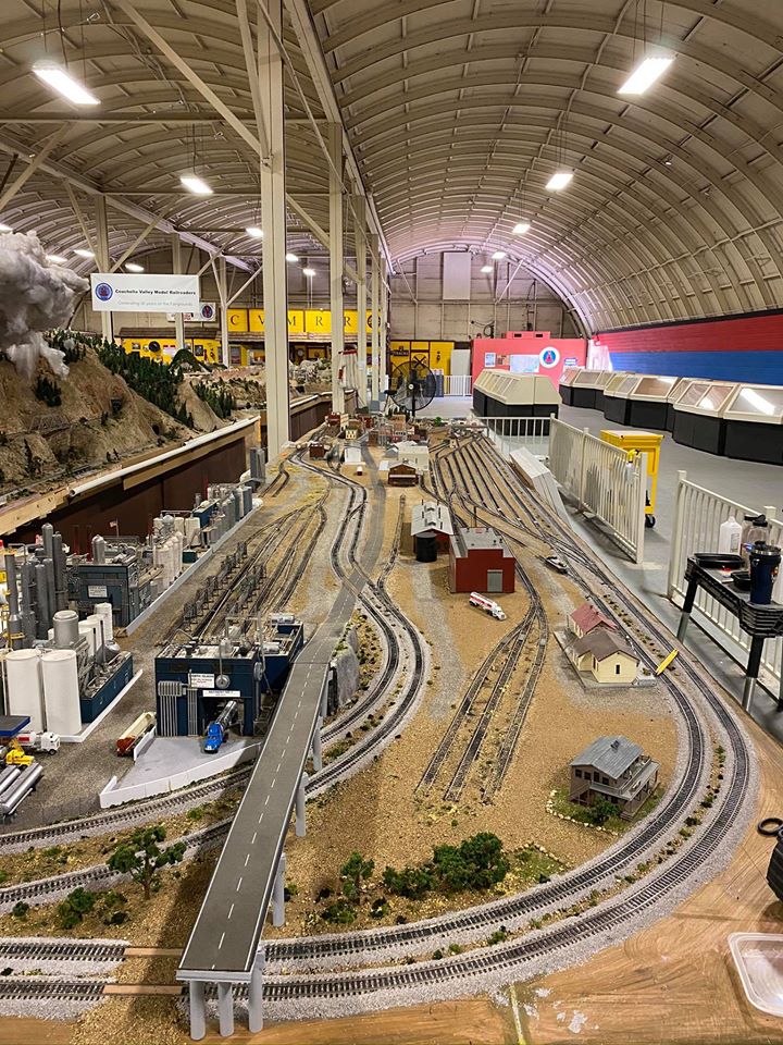 A detailed model train layout featuring winding tracks, industrial buildings, and scenic elements, showcasing a miniature railway environment.