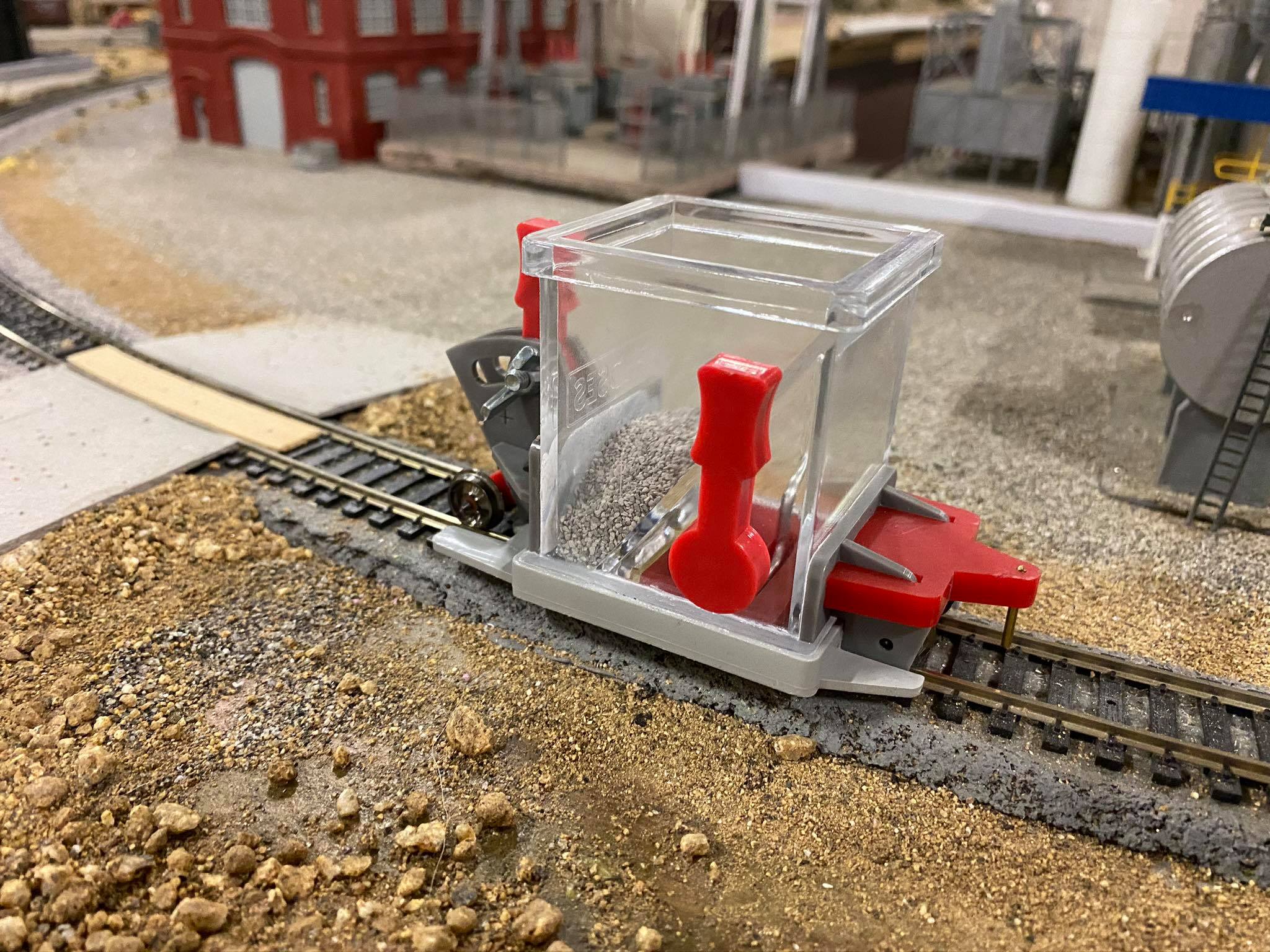 Model railroad maintainer with a clear hopper, positioned on tracks among gravel terrain, highlighting its function in railway upkeep.