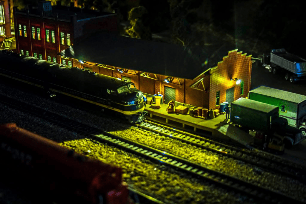 Model train station scene at night, featuring a locomotive beside a brick warehouse. Figures handle cargo under warm illumination, highlighting railway activity.