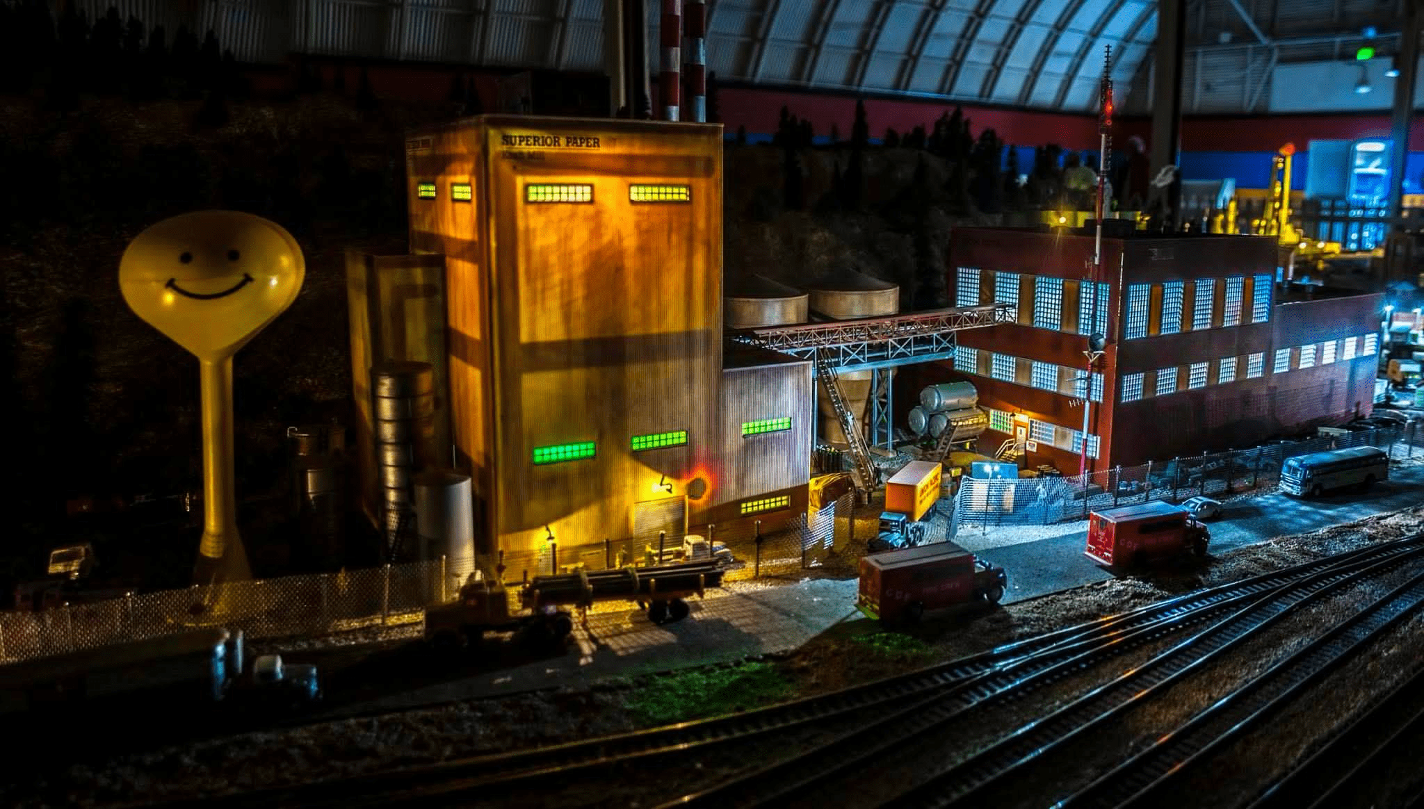 Model train layout depicting an industrial complex with a brightly lit paper factory, storage tanks, and toy vehicles. Ideal for showcasing miniature craftsmanship.